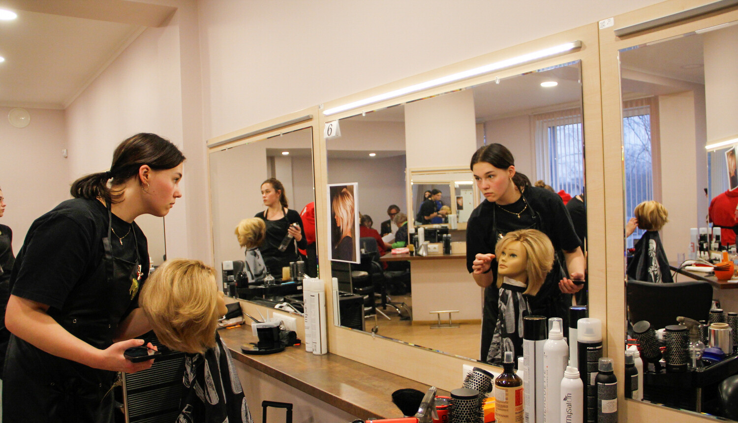 Meitene veido frizūru manekenam un skatās spogulī