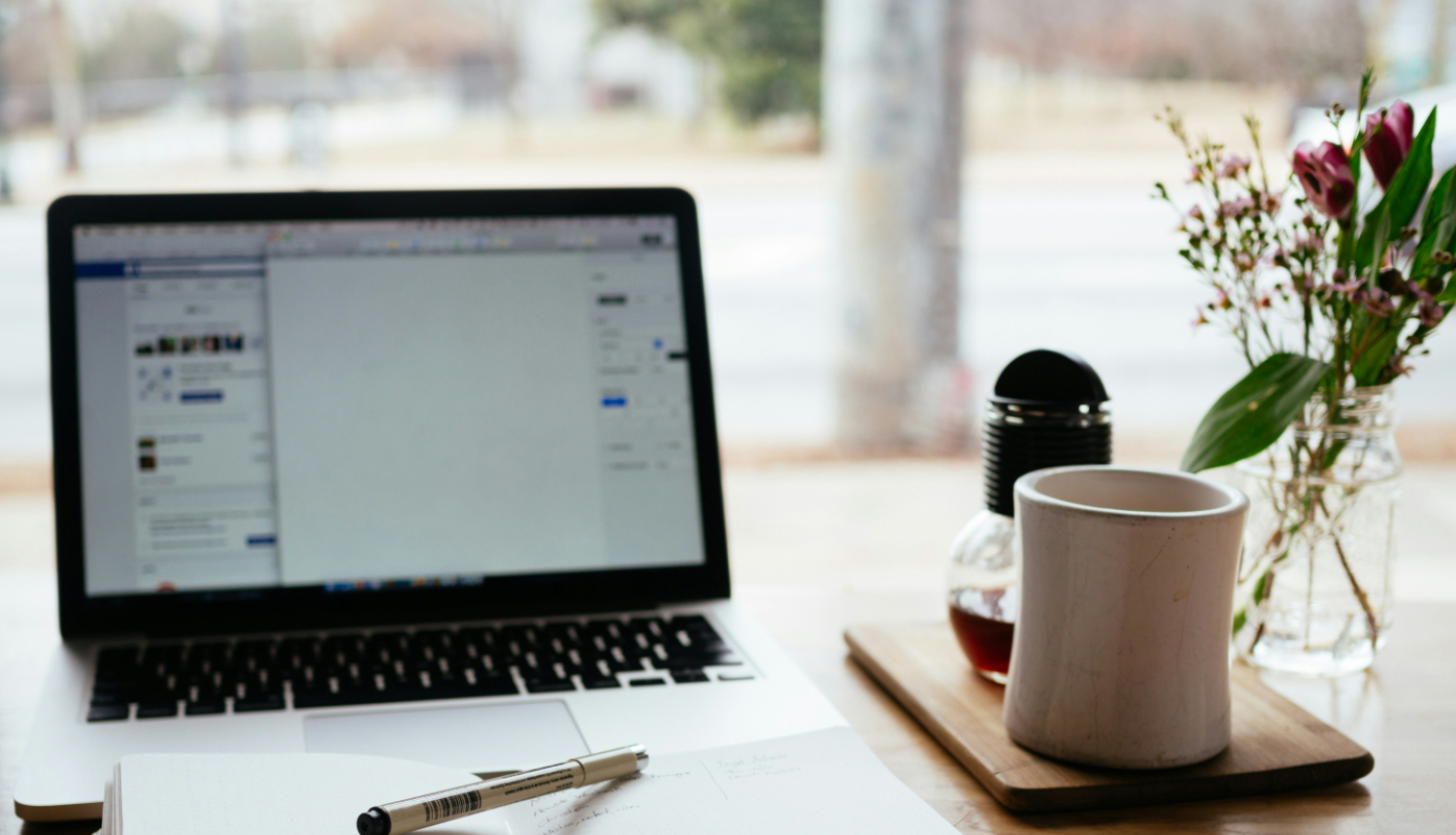 uz galda atvērta pierakstu klade, viršu ir pildspalva, stāv portatīvais dators, blakus krūze un ziedi glāzē