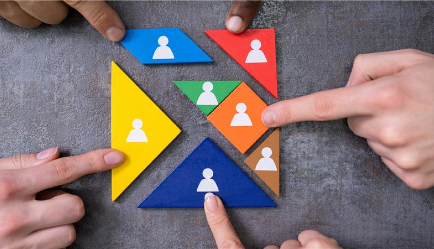 Vairāku cilvēku rokas ar pirktu piebīda klāt krāsainu koka trīstūri, kur attēlota cilvēka ikona