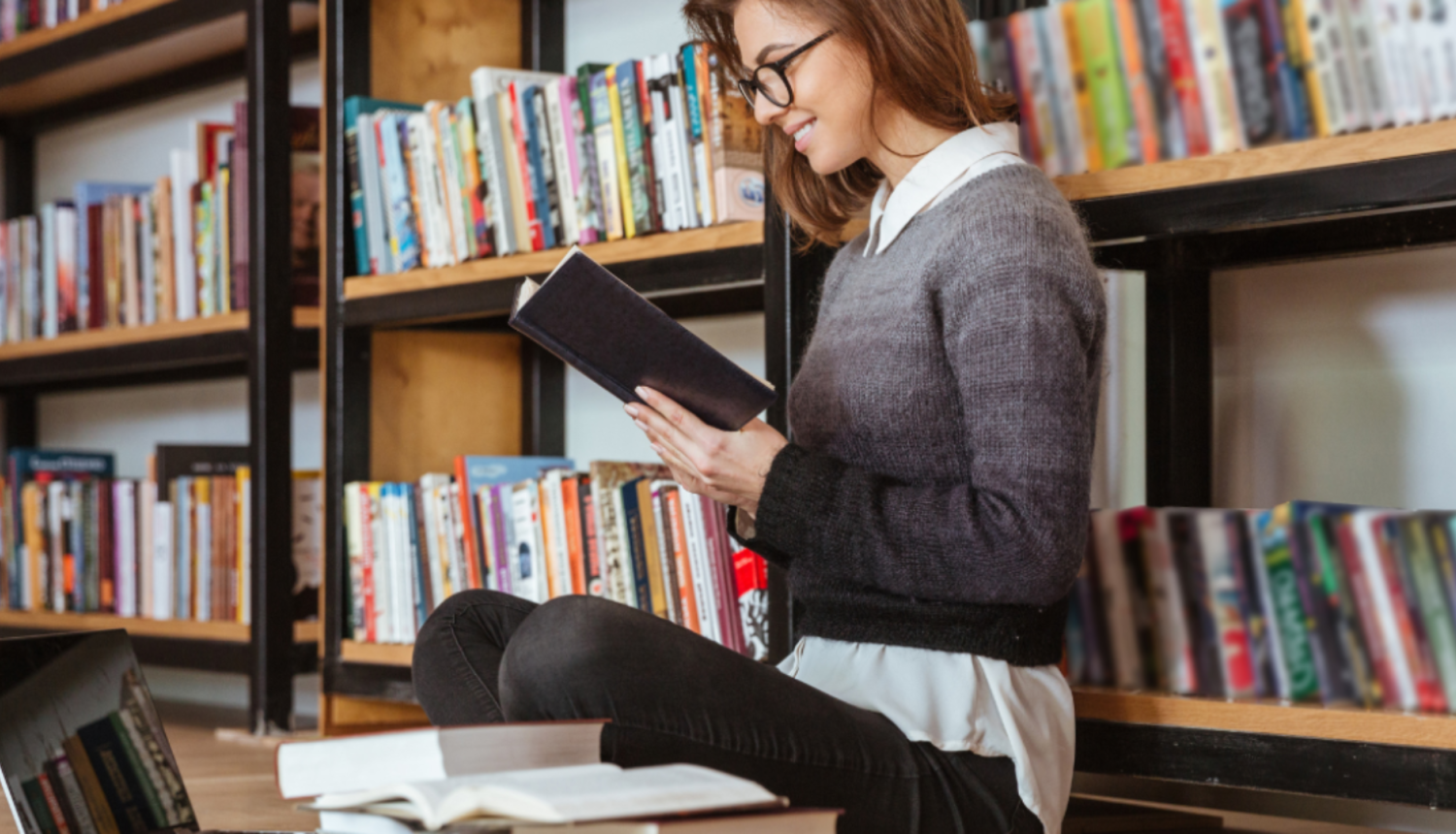 Sieviete sēž un lasa grāmatu pie grāmatu plaukta