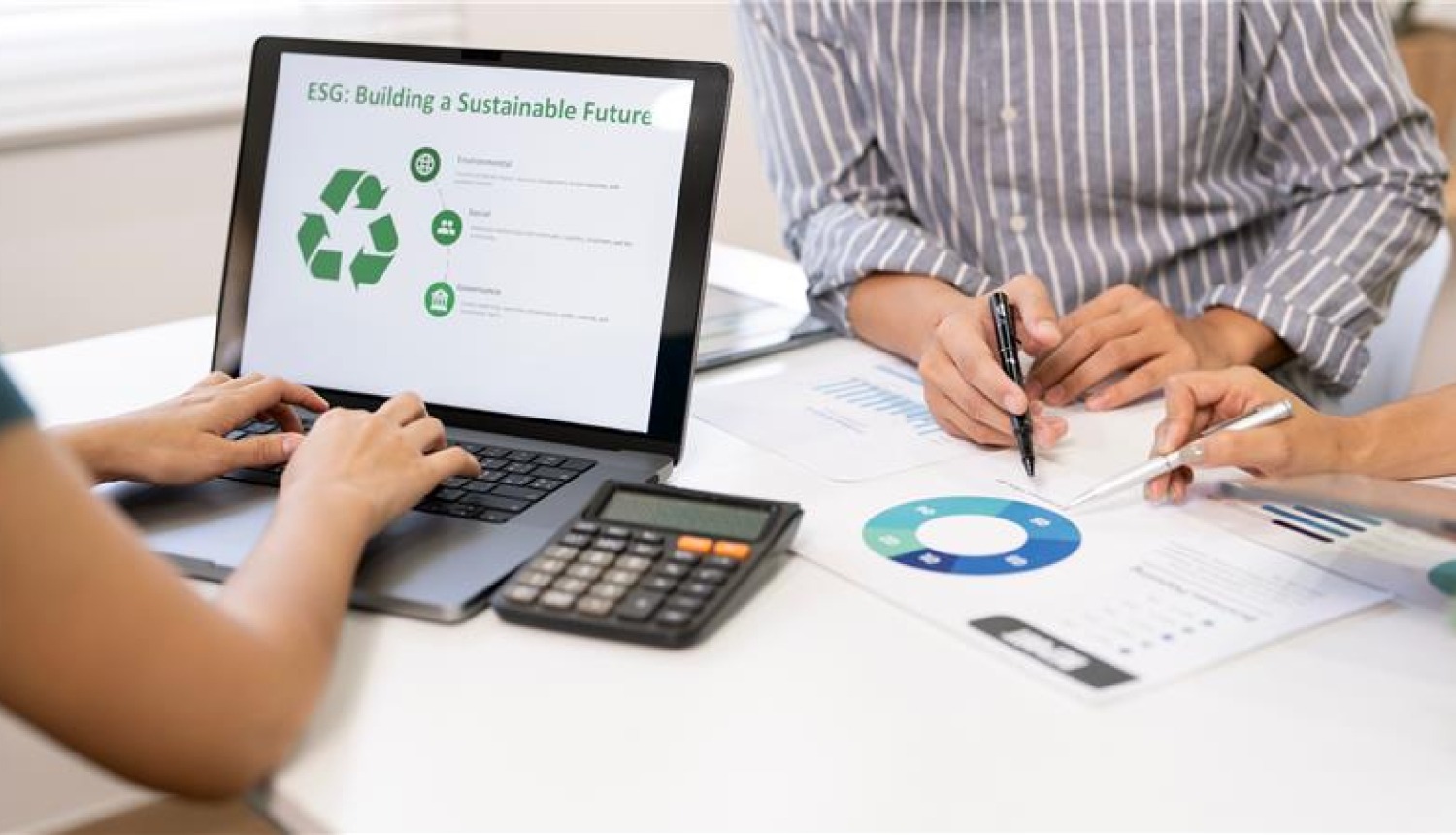 sieviete raksta kaut ko portatīvajā datorā, uz ekrāna viņai ir pārstŗades zaļā zīme, blakus pie galda sēž vīrietis un sieviete un apspriekž shēmu uz papīra