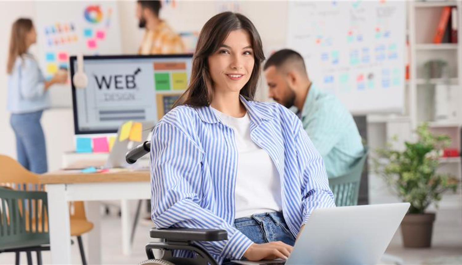 jauna meitene sēž krēslā klēpī tur portatīvo datoru, aizmugurē kāds strādā