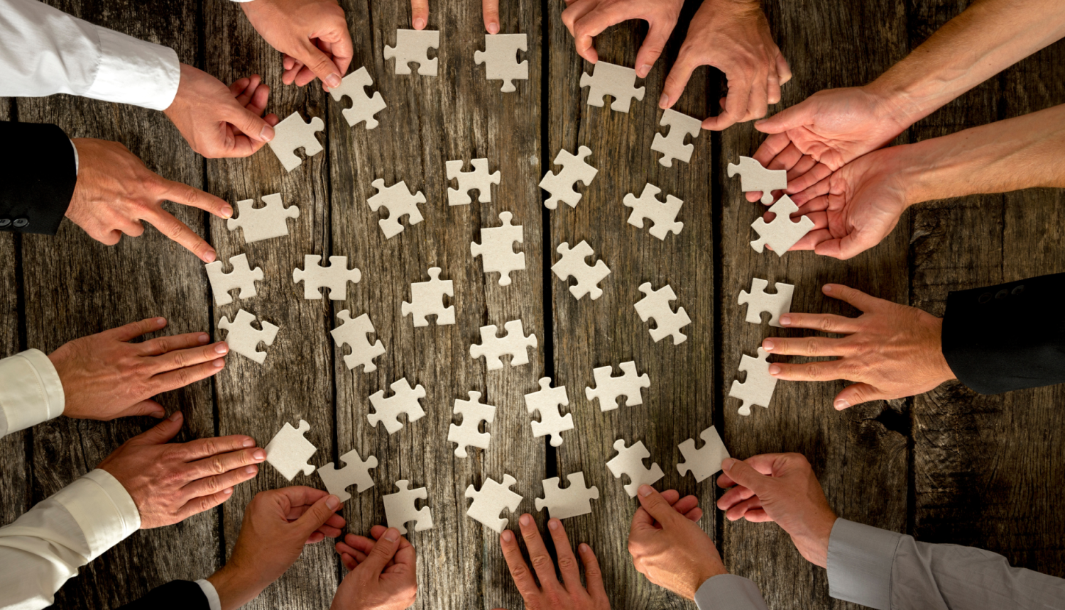 vairāku cilvēku rokas uzliktas uz galda aplī, katra roka mēģina likt kopā puzles gabaliņu