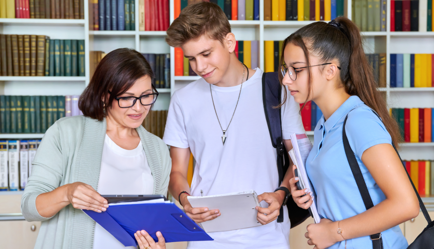 skolotāja rāda kaut ko grāmatā diviem studentiem