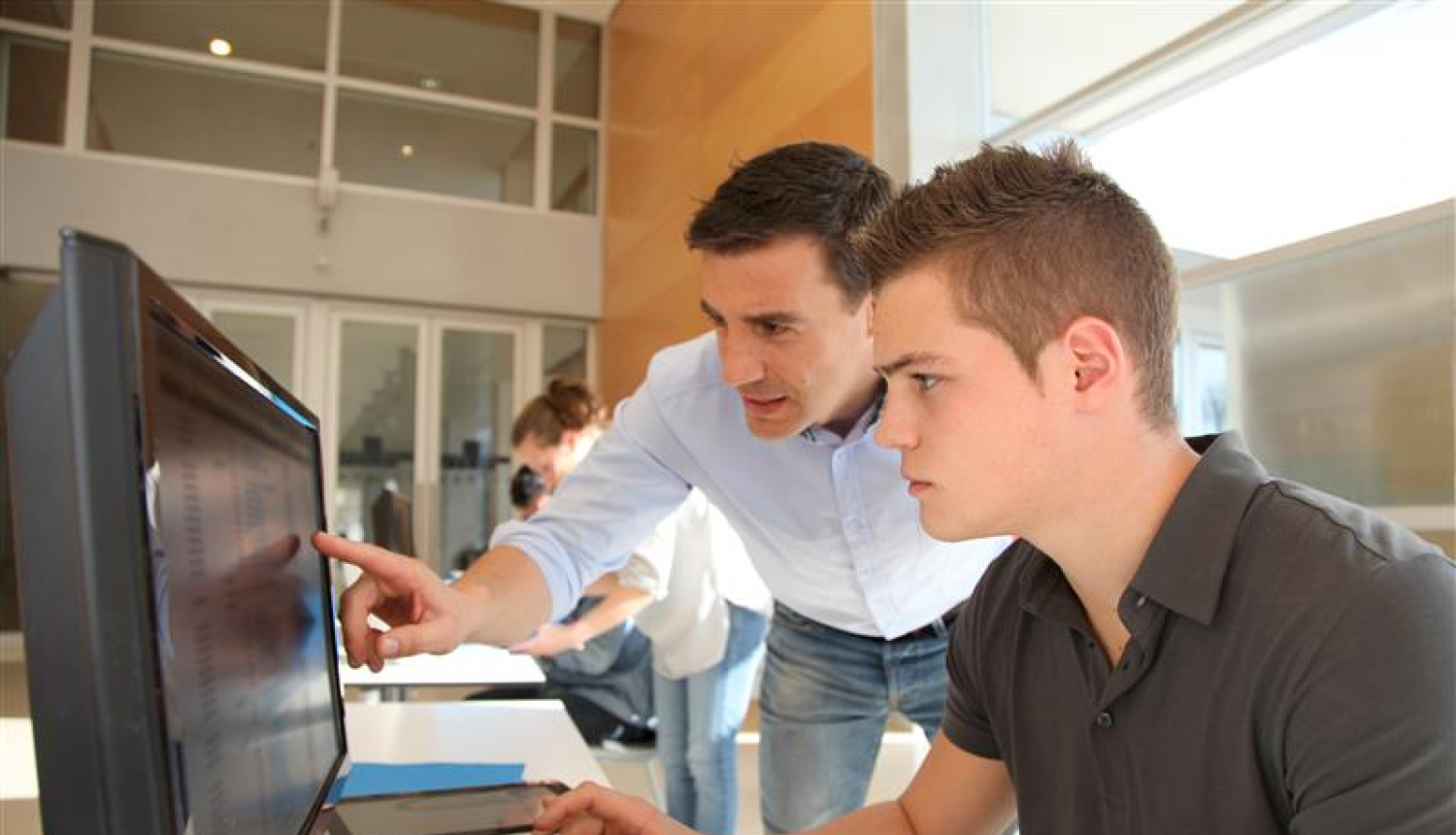 jaunietis sēž pie datora klasē, blakus stāvošais vīrietis rāda kaut ko viņam monitorā 