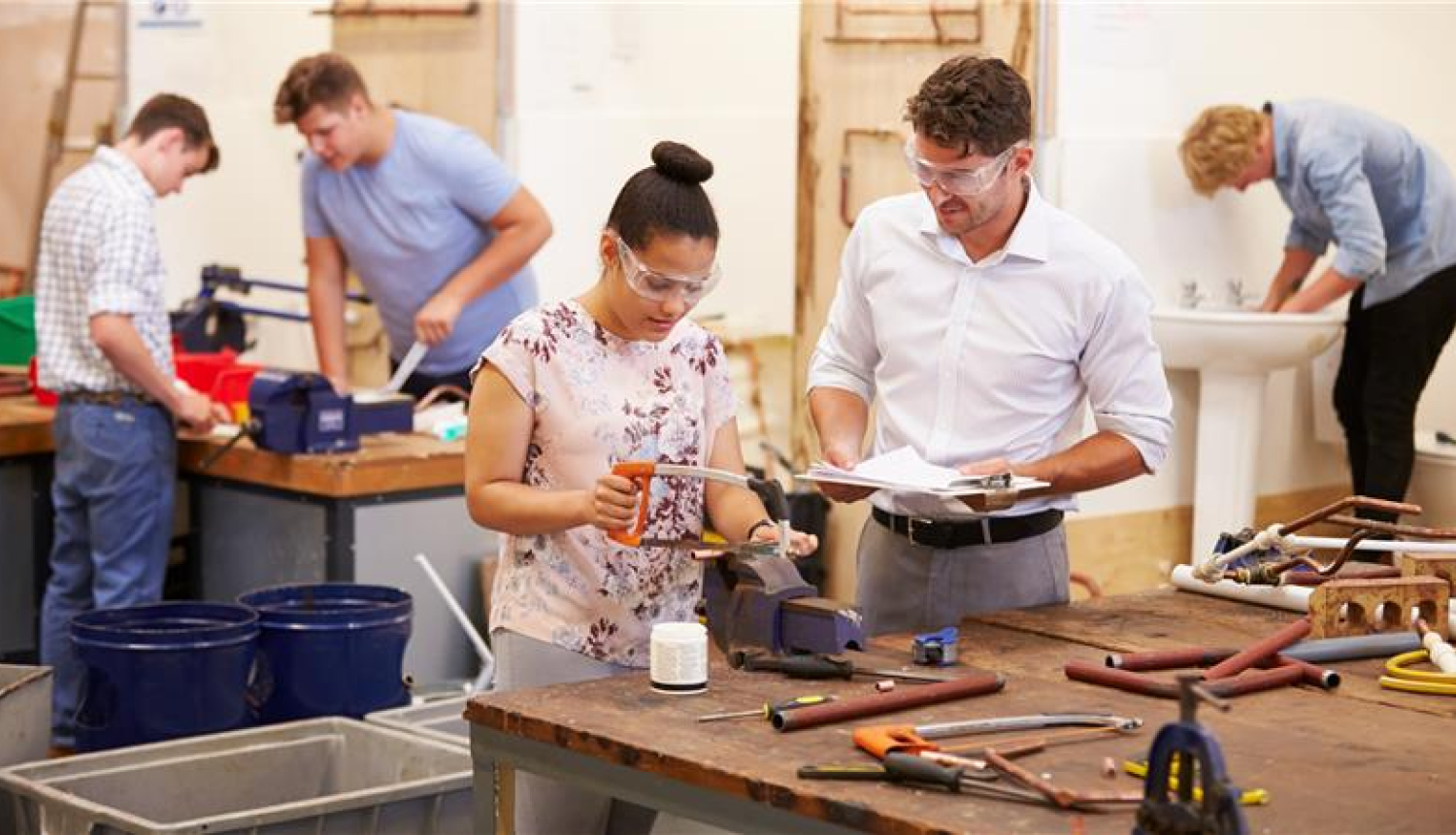 sieviete strādā ar zāģi vīrietis skatās kā viņa strādā, aizmugurē'jā plānā arī strādā trīs vīrieši ar žāģi