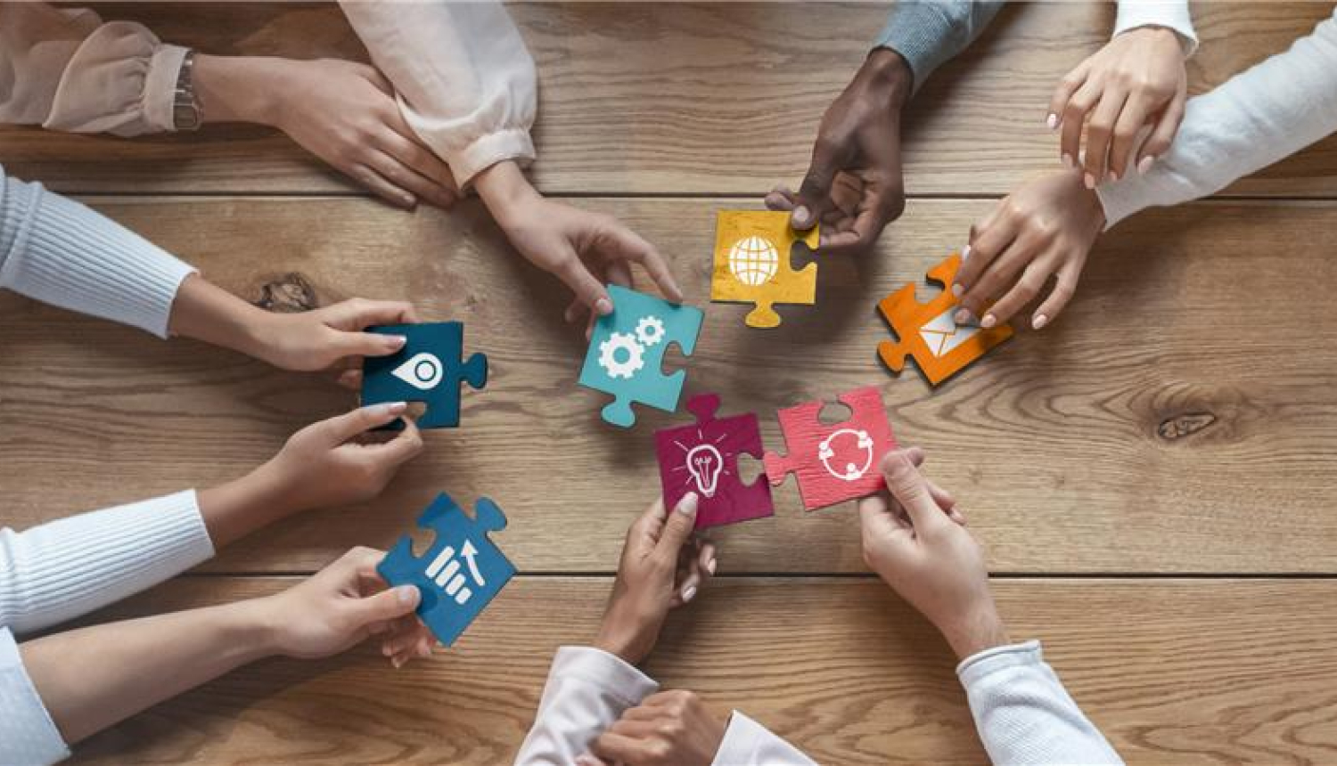 vairākas cilvēku rokas virs galda tur lielus puzles kauliņus un mēģina tos salikt kopā