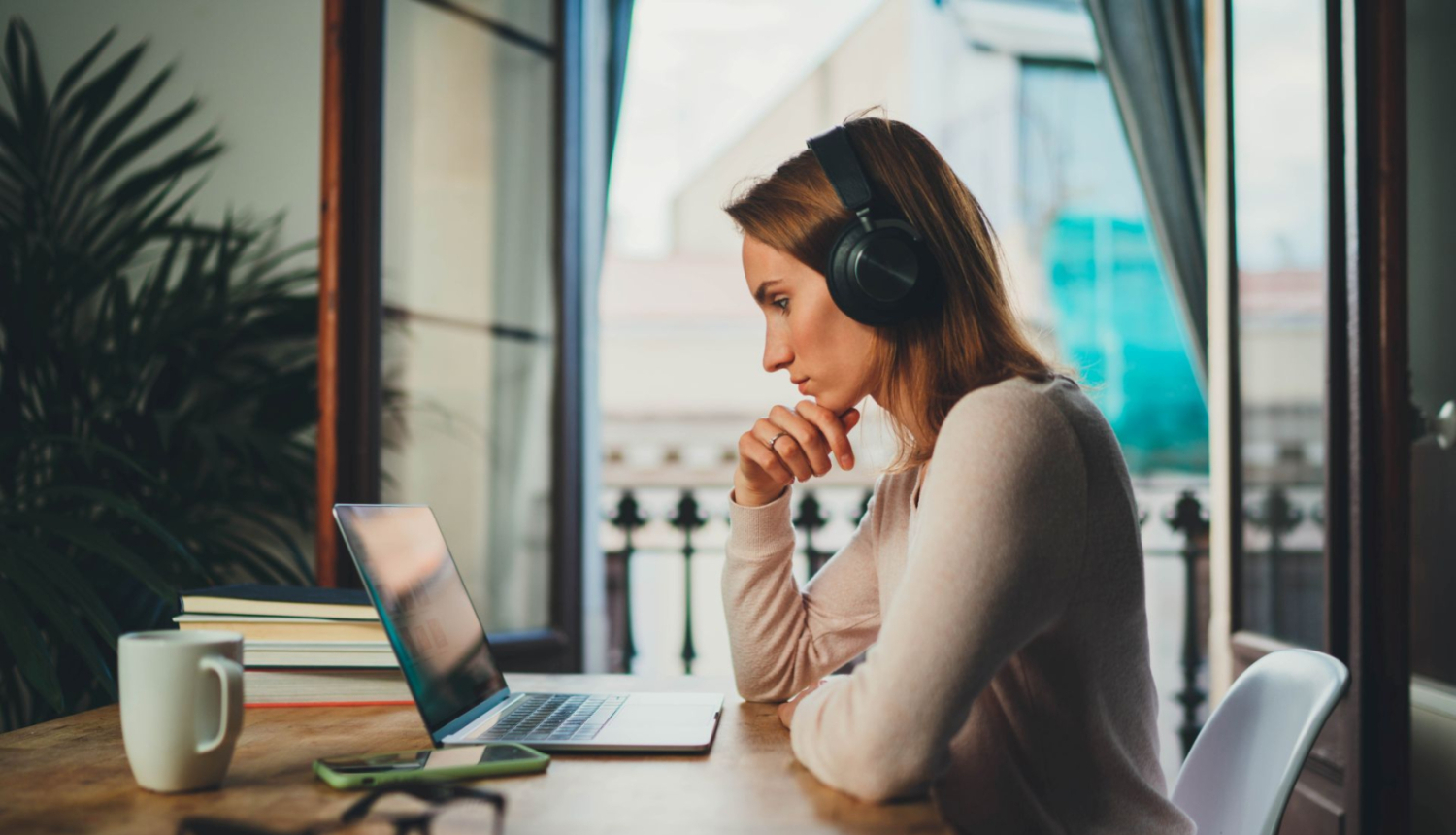 jauna sieviete sež pie galda ar austiņām uz galvas, priekšā viņai uz galda atveērts portatīvais dators