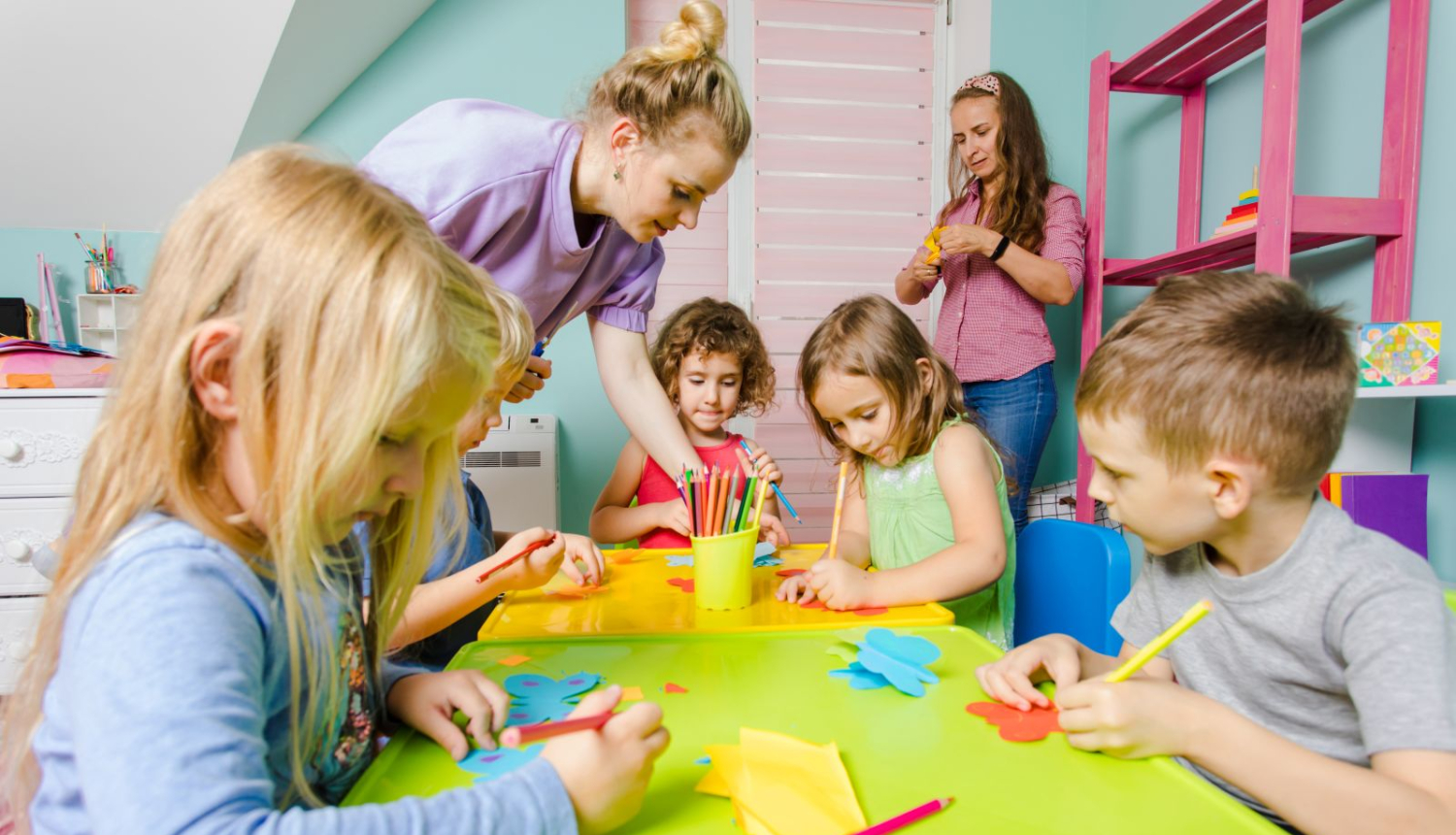 bērni zīmē pie  galda pirmskolas izglītības iestādē blakus audzinātāja palīdz bērniem