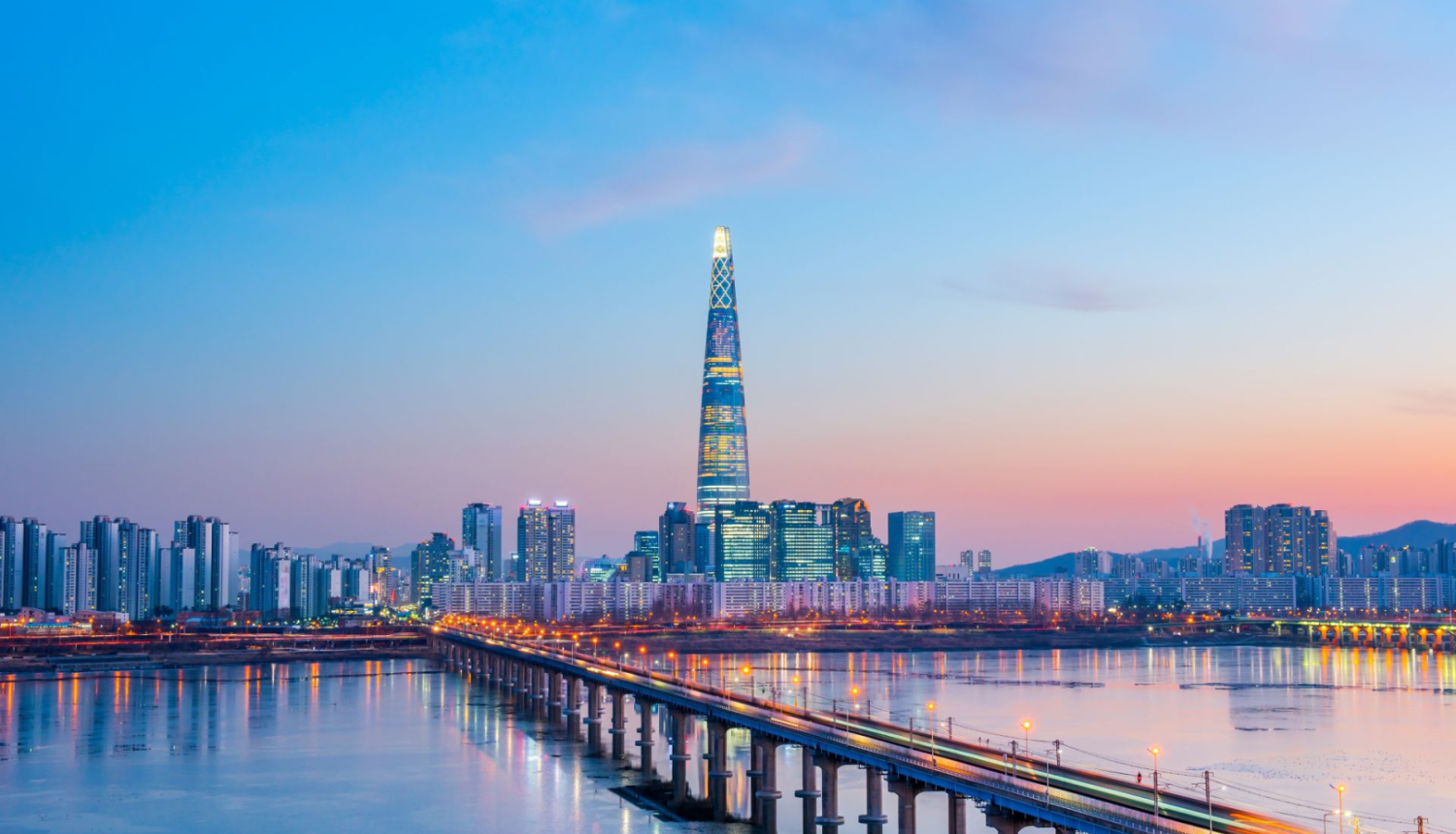attēlā skats uz Korejas galvaspilsētu, priekšā tilts pāri upei, aizmugurējā plānā augšceltne, saulriets