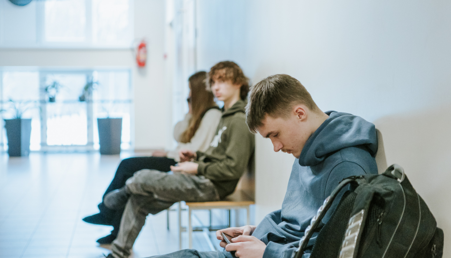 skolēni ar telefonu rokās sēž uz krēsla skolas gaitenī 