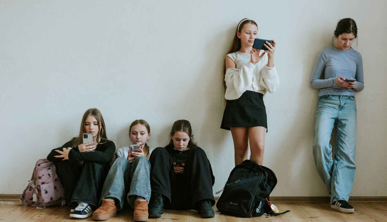 pusaudži sēž uz grīdas un stāv pie sienas visi skatās telefonos