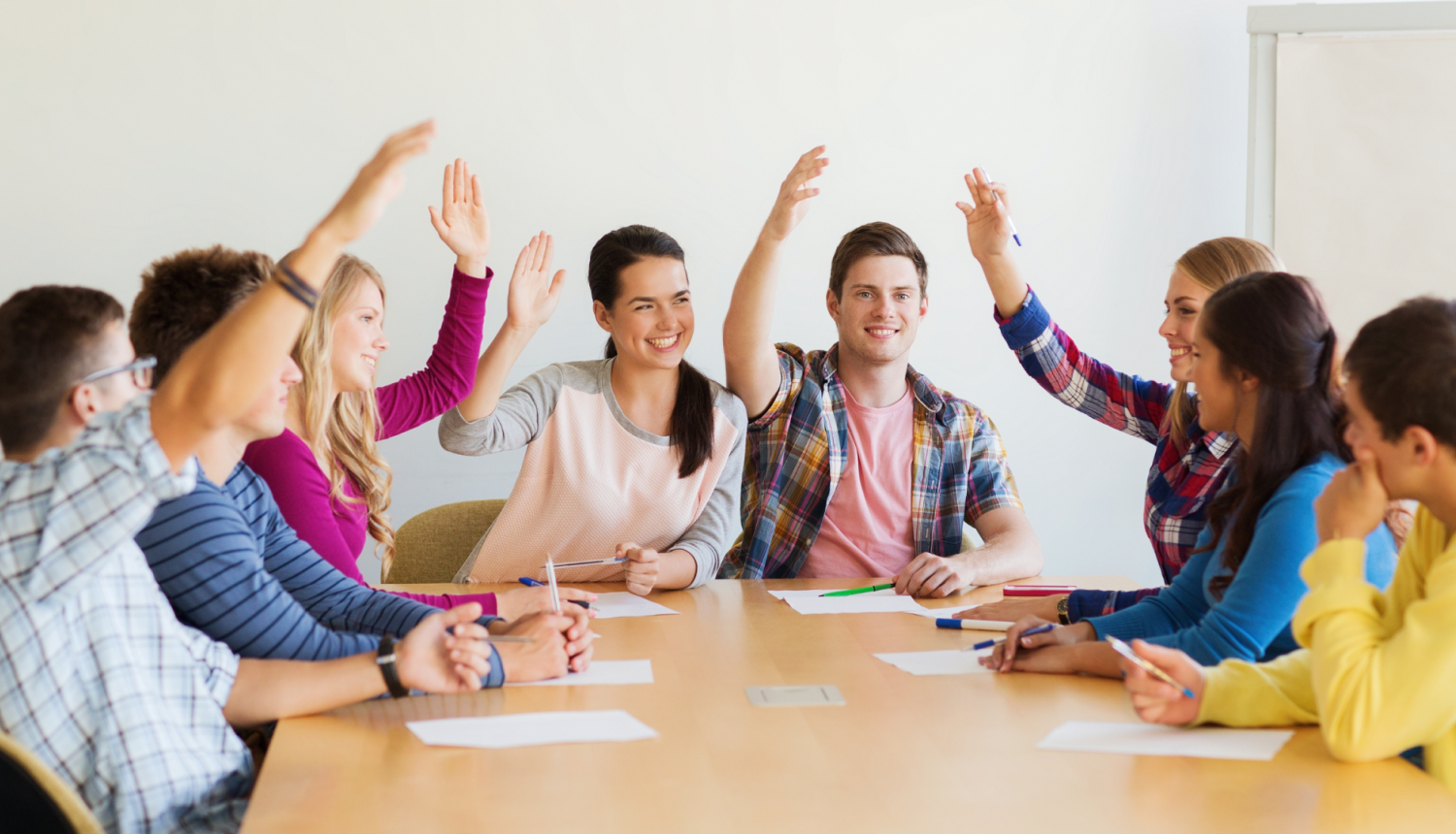 skolēnu grupa sēž pie galda un paceļ rokas it kā par kaut ko kopīgi balsotu
