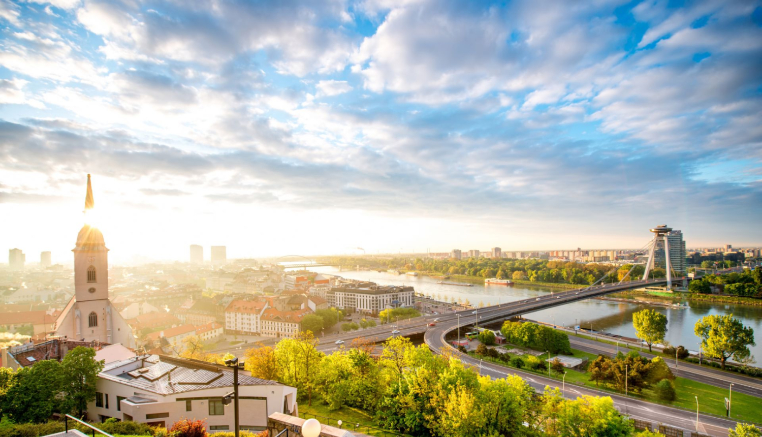 Attēlā skats uz Slovākijas galvaspilsētu, pa kreisi baznīcas tornis, pa visu upe, fonā spoža saule