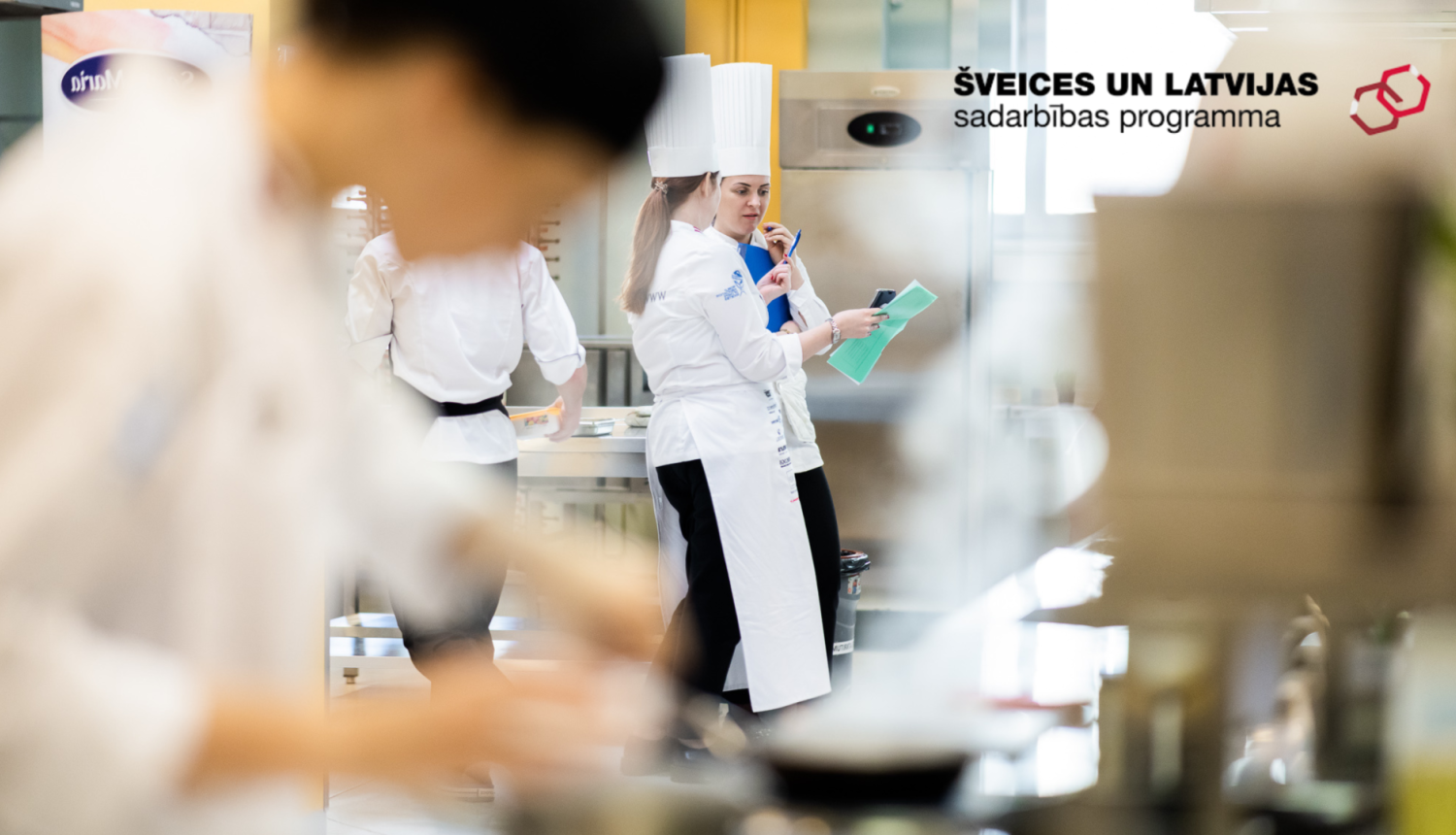 Māceklis pavārs priekšplānā gatavo aizmugurējā plānā pavāru pasniedzējs sniedz norādījumus