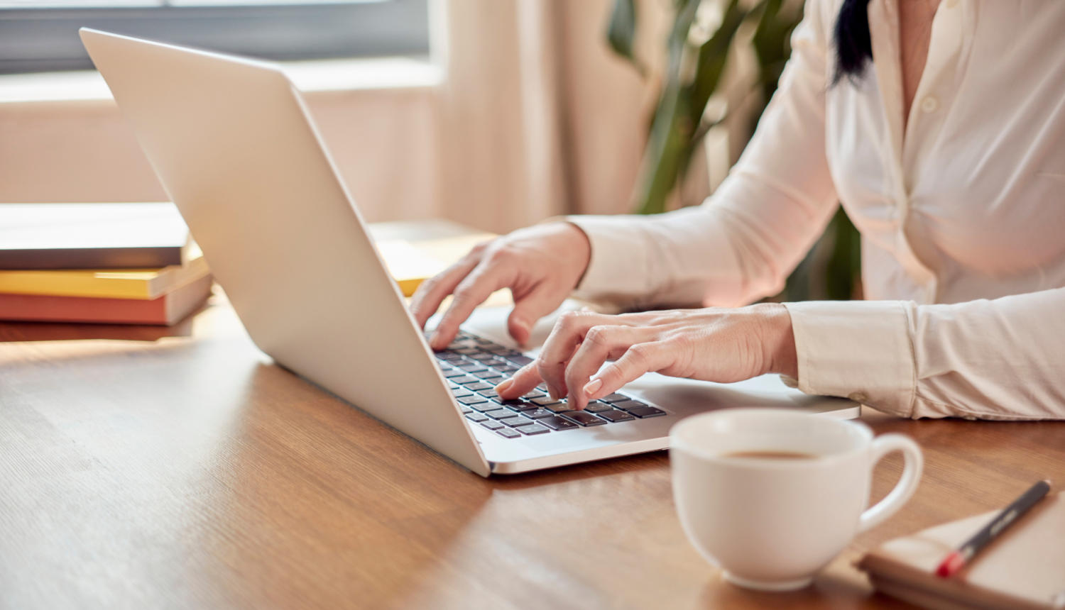 attēlā sieviete sēž pie portatīvā datora raksta uz klaviaraturas, uz galda stāv krūze