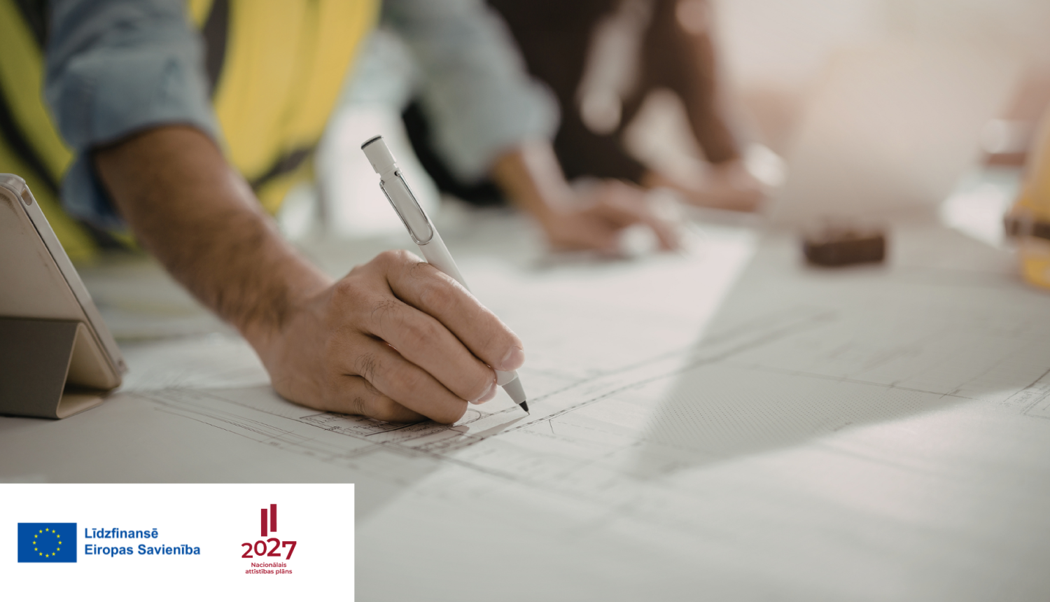 vīrietis atstarojošā darba vestē atzīmē kaut ko ar pildspalvu uz lielā būvdarbu rasējuma, kas stāv uz galda