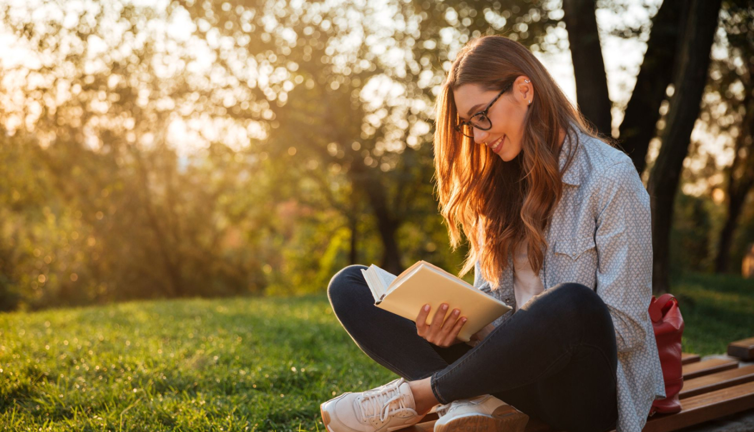 studente ar brillēm sēž parkā uz soliņa un lasa grāmatu