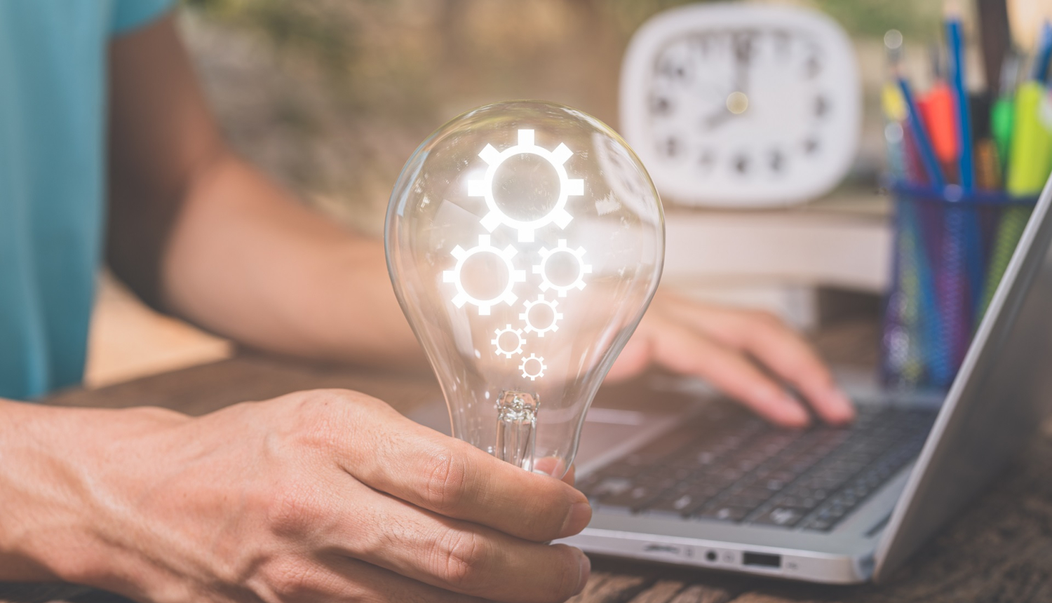 Vīrieša roka, atstutēta pret galdu, tur ieslēgtu spuldzi blakus datoram