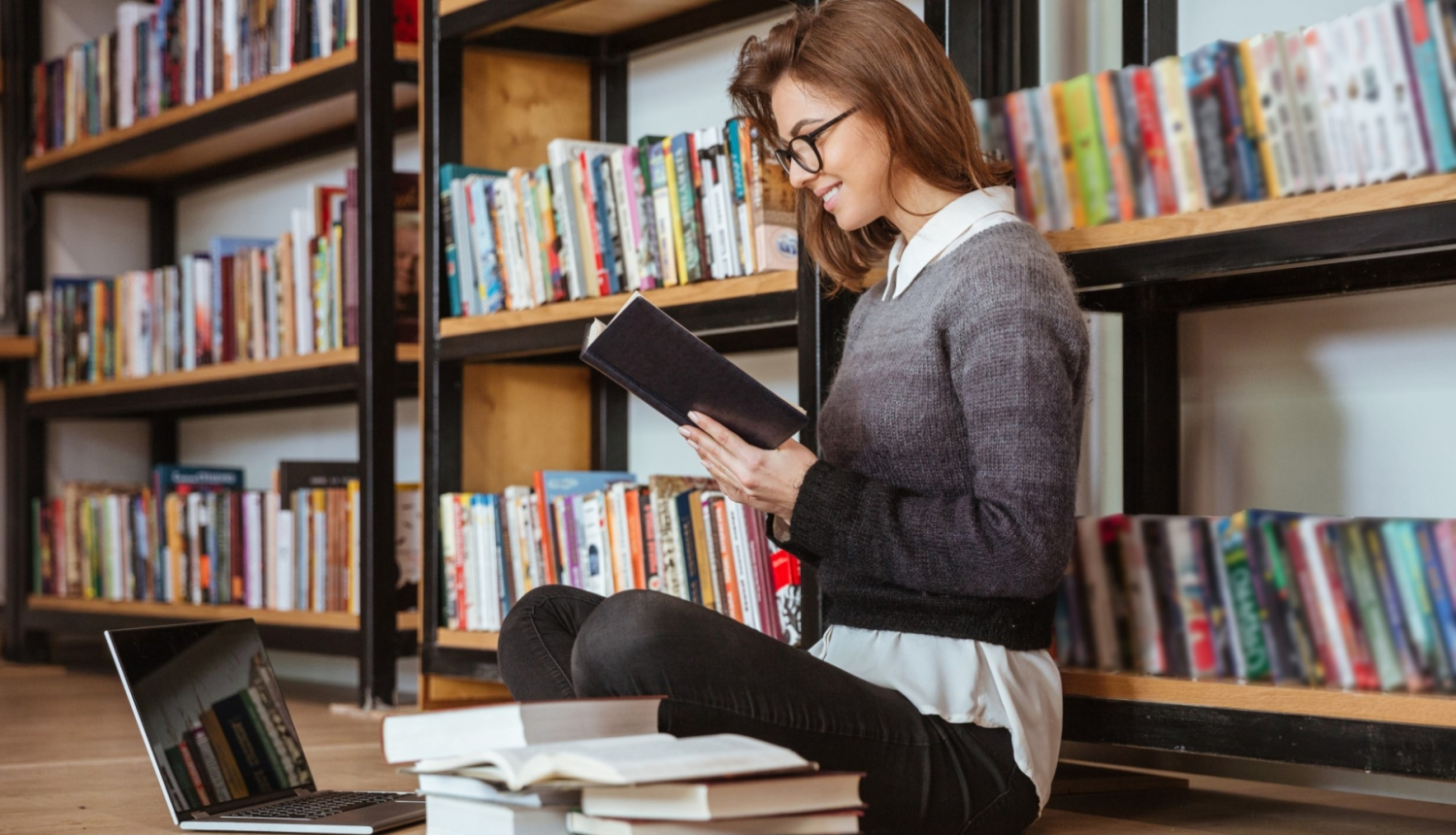 jauna sieviete ar brillēm sēž uz grīdas ar grāmatu rokās biliotēkā