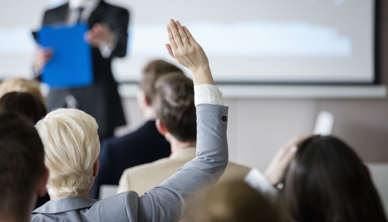 sieviete ceļ augšā roku semināru zālē priekšā stāv vīrietis uzvalkā pie tāfeles