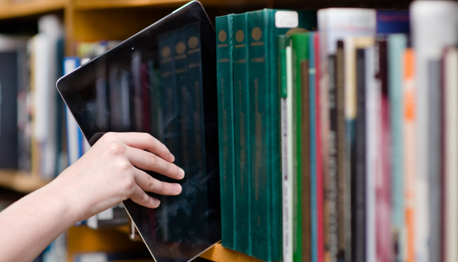 cilvēka roka stiepjas ar planšeti to ievietot grāmatu plauktā bibliotēkā