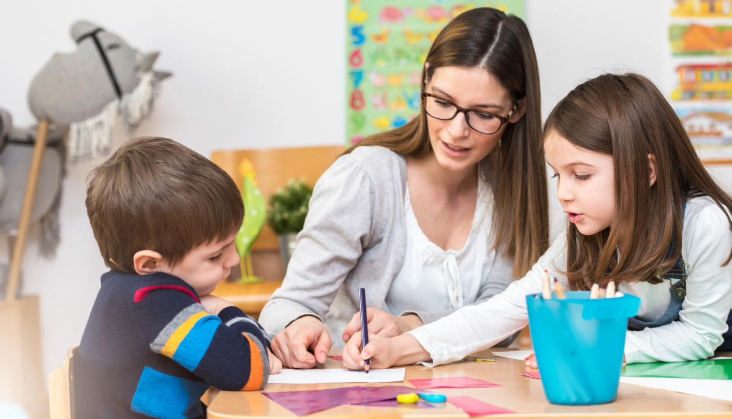 skolotāja zīmē kopā ar puisīti un meitenīti pie galda bērnudārzā