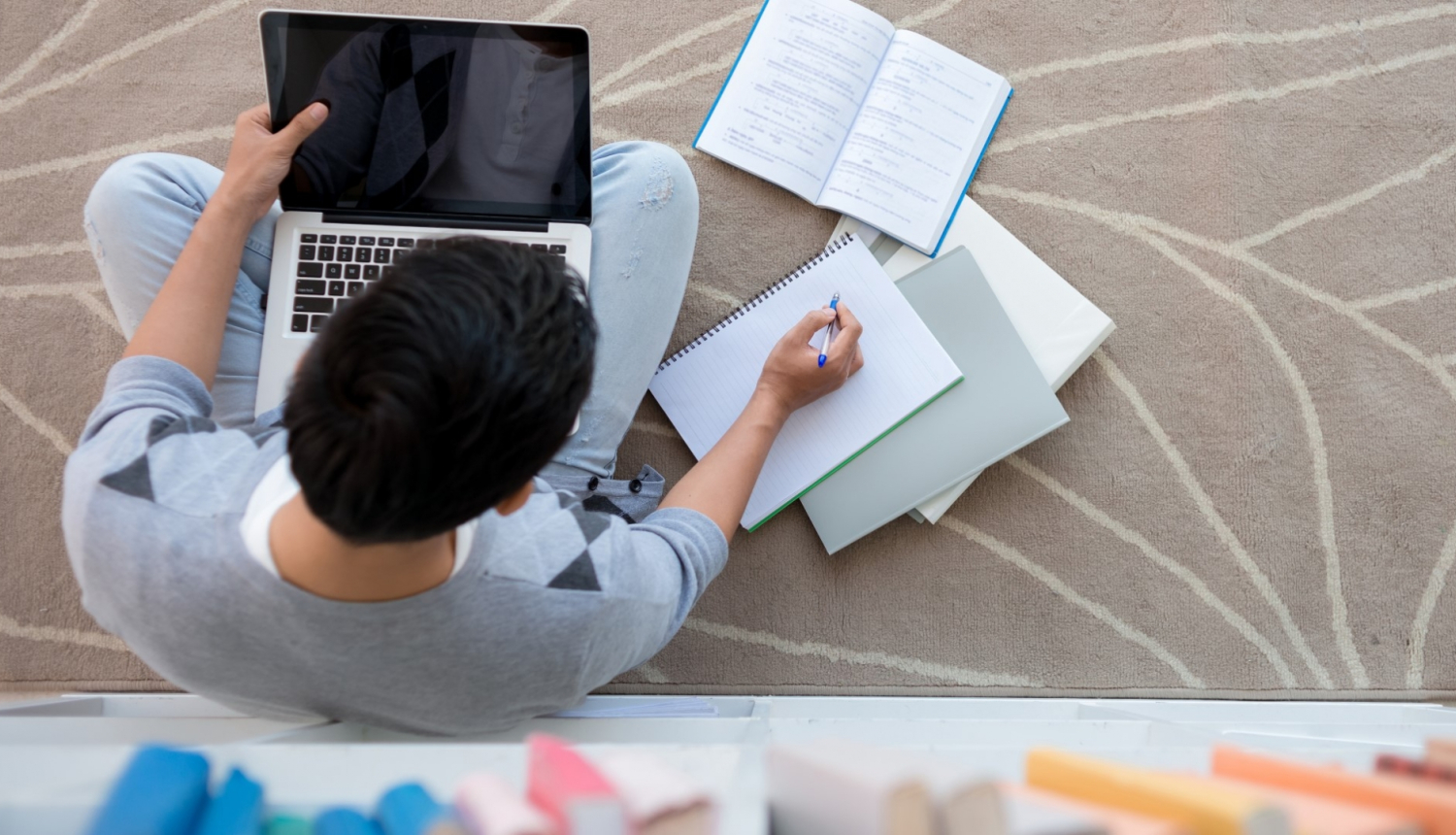 jauns vīrietis sēž uz grīdas bibliotēkā kaut ko pieraksta kladē un uz ceļiem tur klēpjdatoru