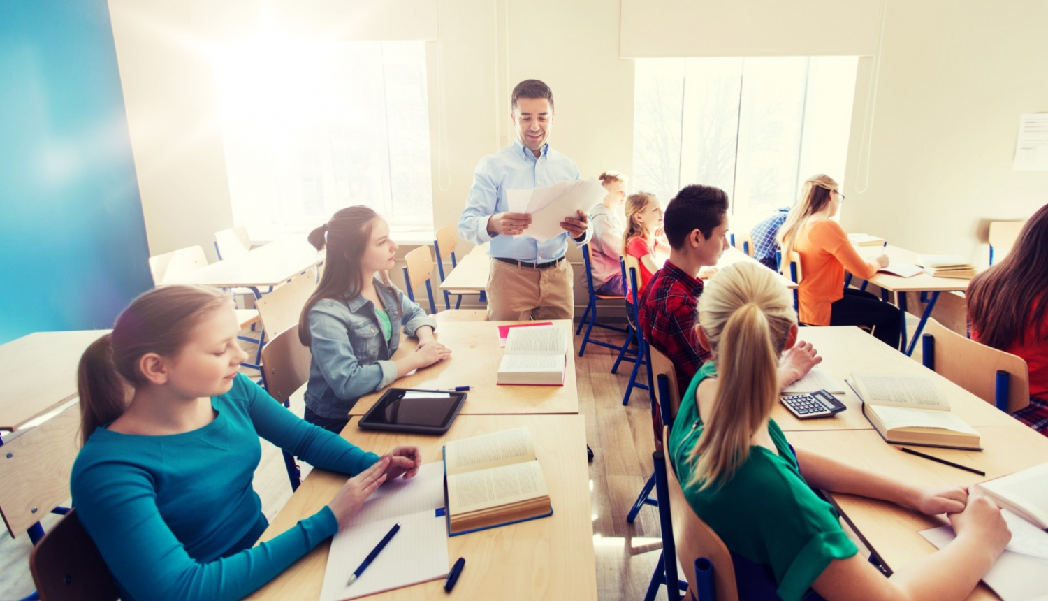 skolēni sēž klasē blakus stāv smaidīgs skolotājs ar darba lapām