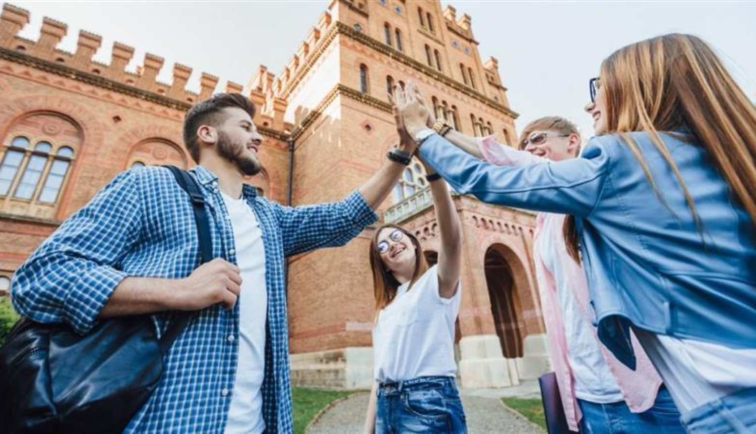 Jaunieši studenti sasit priecīgi plaukstas, fonā universitātes ēka