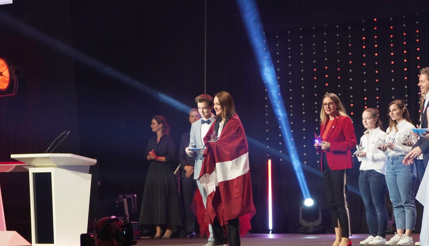 Meitene uz skatuves ar Latvijas karogu ap pleciem