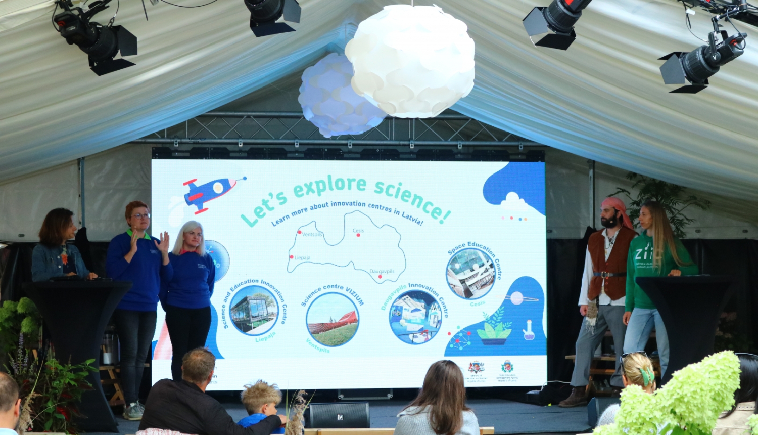 Cilvēki uz skatuves zem nojumes stāsta par inovāciju centriem