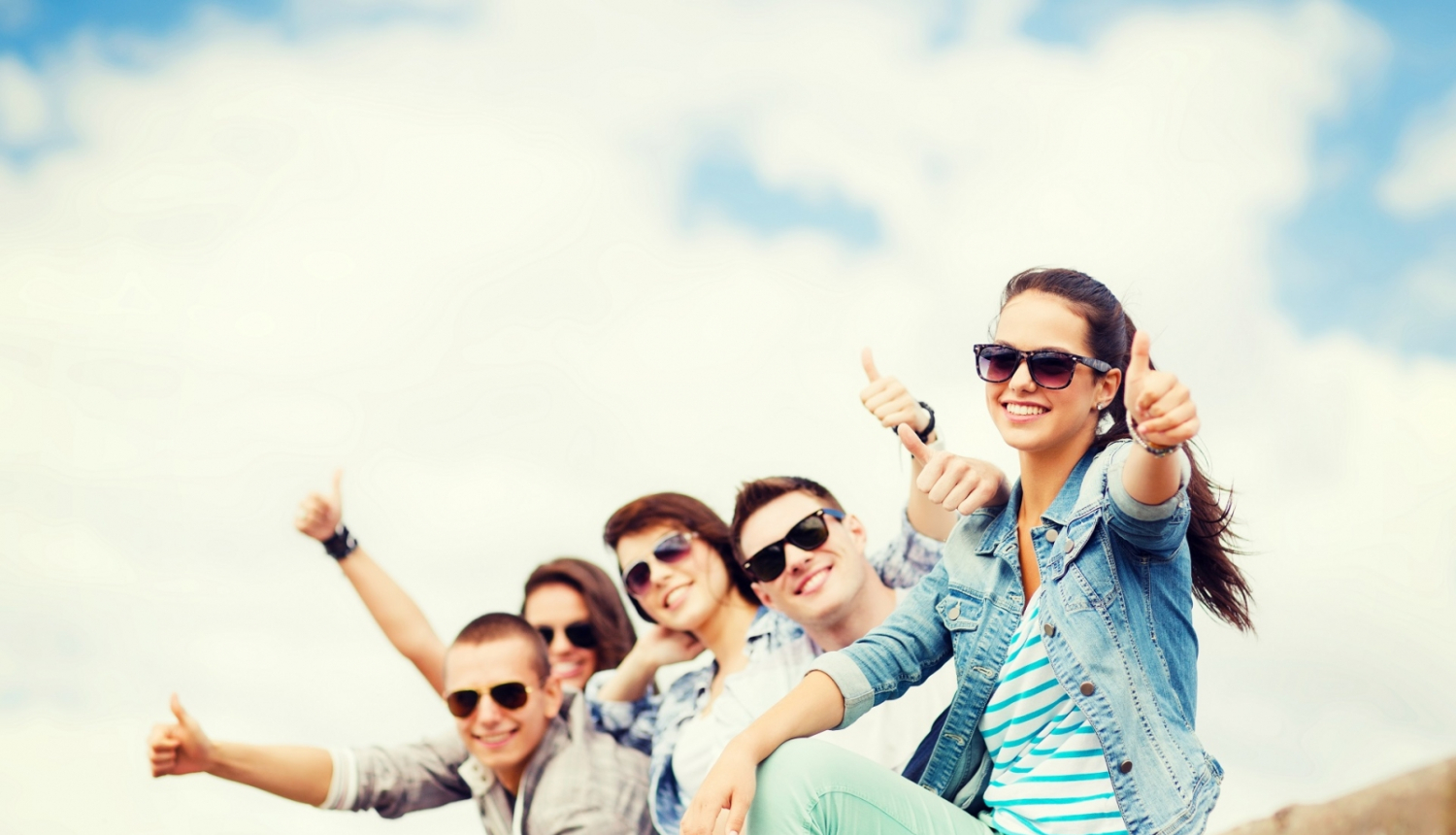 Jaunieši saulesbrillēs sēž uz zeme, smaida un rāda lielo īkšķi