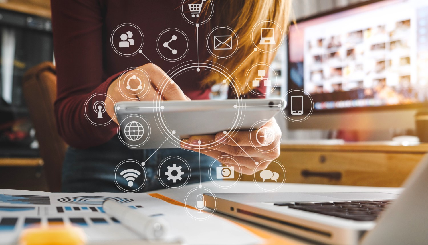 Darba galds ar klēpjdatoru, sieviete tur rokās planšeti, kurā attēlotas ikonas