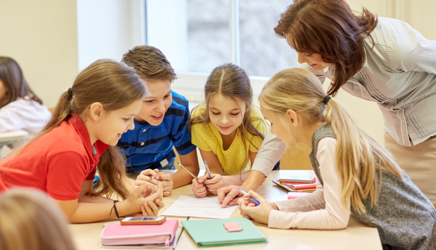 vairāki skolēni sēž grupā pie galda skolotāja stāv blakus un kaut ko norāda viņiem