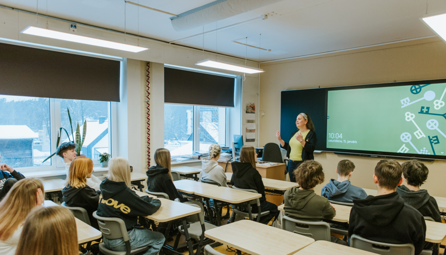 skolēni sēž pie soliem klasē, klases priekšā stāv skolotāja pie interaktīvās tāfeles 