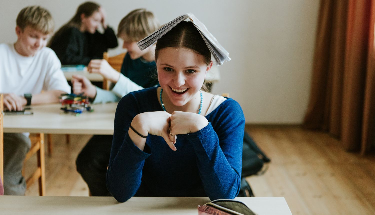 meitene skolniece sēž skolas solā un smaida, uz galvas viņai ulikta atvērta grāmata; aizmugurējā plānā sēž skolas solā citi skolēni