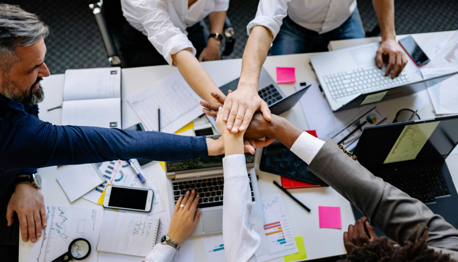 vairāki cilvēki sēž pie viena darba galda pa apli un pa vidu galdam sakruastoja rokas, uzliekot vienu otram virsū; uz galda ir dažādas pierakstu lapas, telefons, dators