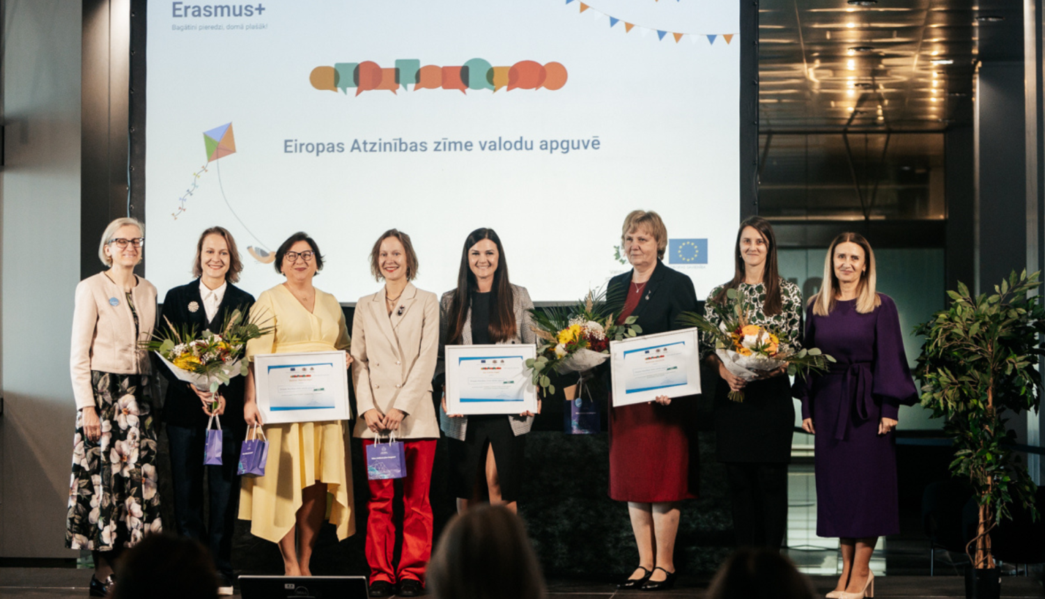 Sievietes stāv uz skatuves ar ziediem rokās