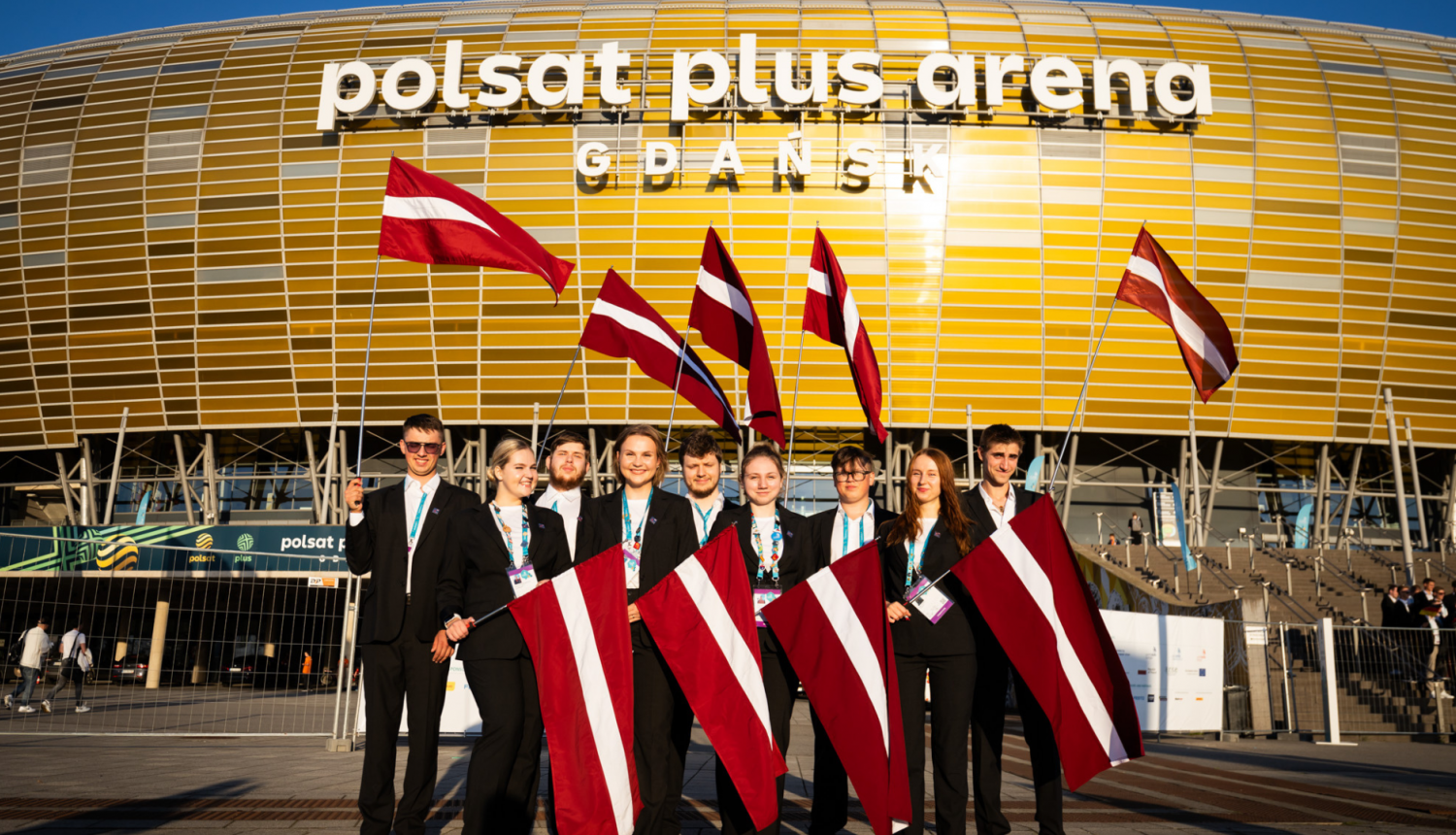 Jaunieši ar Latvijas karogiem rokās stāv priekšā arēnai