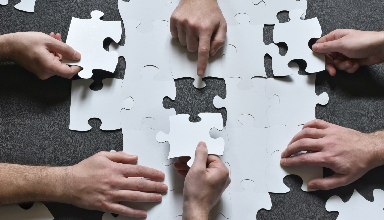 vairākas rokas uz galda liek kopa puzles gabaliņus