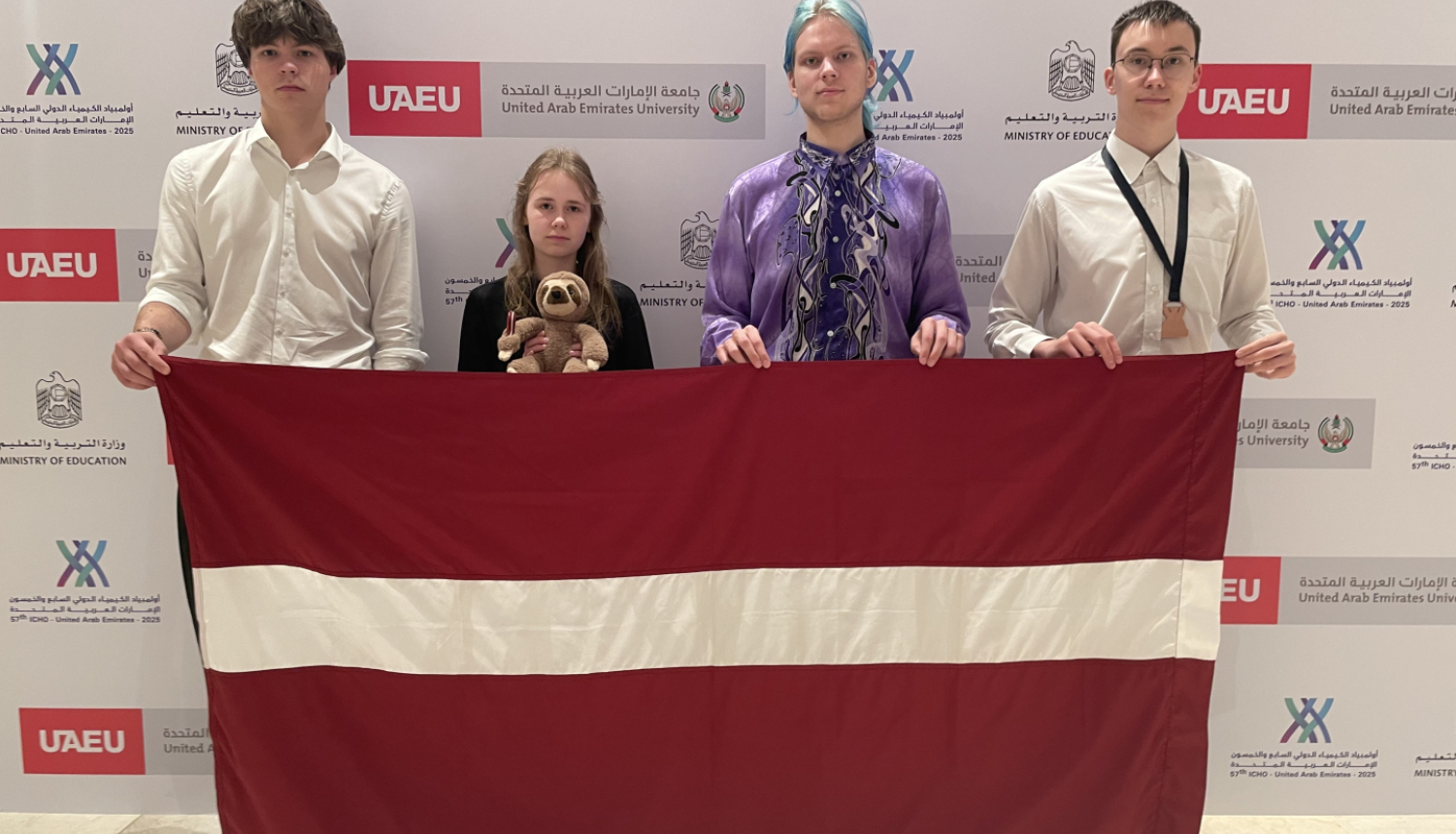 Latvijas komandas dalībnieki rokās ar LV karogu ķīmijas olimpiādē 2025 Dubaijā