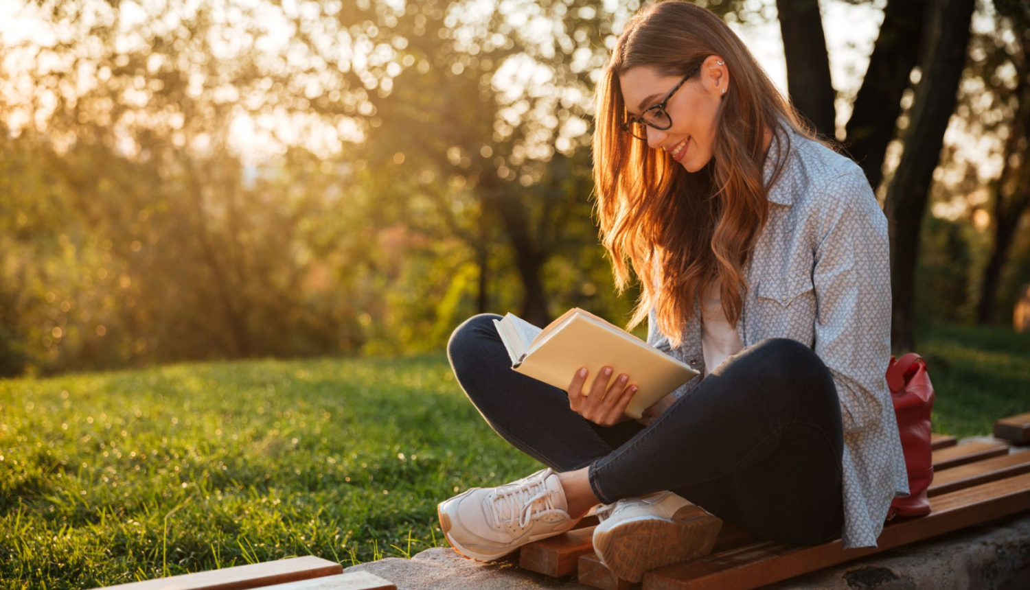 studente ar brillēm sēž parkā uz soliņa un lasa grāmatu