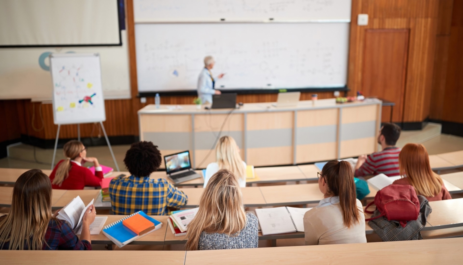 Augstskolas auditorijā lektors stāv pie lielas tāfeles, auditorijā sēž studenti un klausās