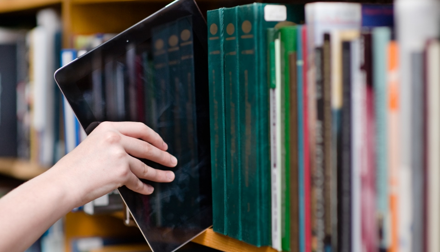 cilvēka roka stiepjas ar planšeti to ievietot grāmatu plauktā bibliotēkā