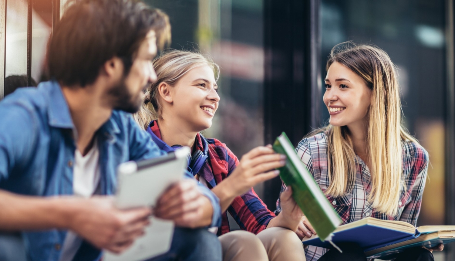 trīs studenti sēž uz trepēm ar grāmatām